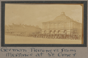Eine Gruppe deutscher Gefangener aus Messines vor einem Gebäude unter einem klaren blauen Himmel, mit Text am unteren Bildrand.