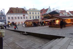 Ein geschäftiger Weihnachtsmarkt in Tallinn, Estland, mit Menschen um geschmückte Stände, festlicher Beleuchtung, bewölktem Himmel, Gebäuden im Hintergrund, Treppen nach oben und Töpfen mit Pflanzen herum.