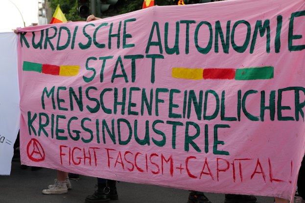Gruppe von Menschen mit einer pinken Fahne, auf der "Kurdische Autonomie Statt Menschenfeindlicher Kriegsindustrie Fight Fascism Capital" steht, auf einer Straße mit Ampeln, Flaggen, Bäumen, Gebäuden und einem klaren blauen Himmel im Hintergrund.