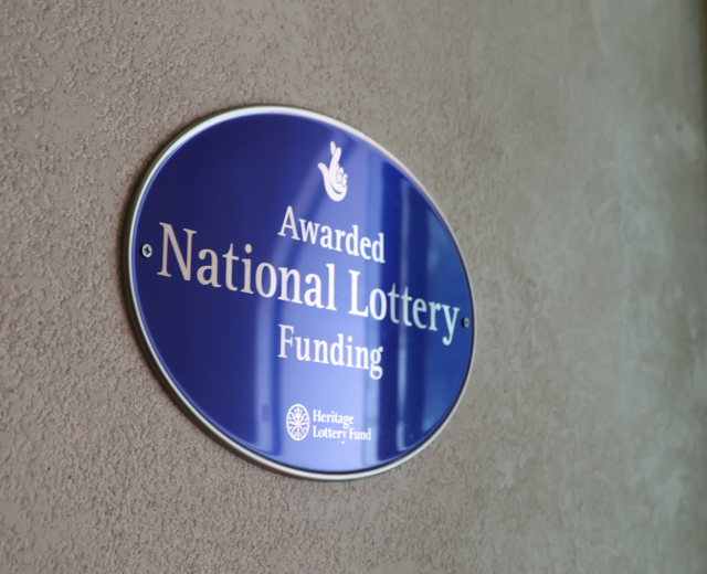 Blaues rechteckiges Schild mit weißem Hintergrund und schwarzer fettgedruckter Schrift, die "National Lottery Funding" lautet.