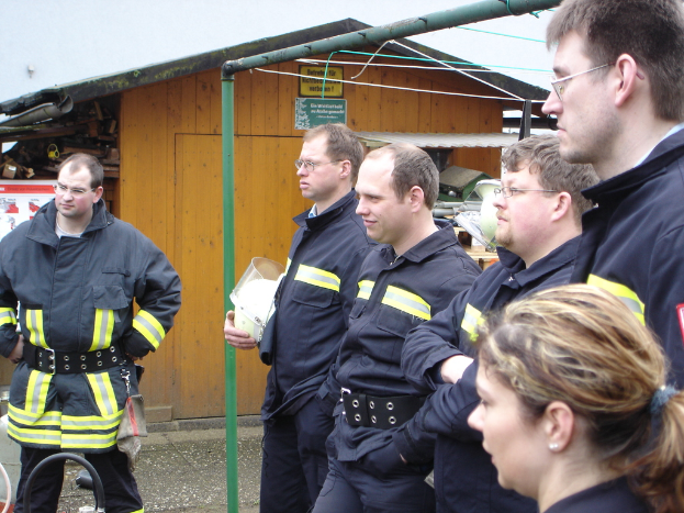 Eine Gruppe von Männern in blauen Feuerwehreinsatzuniformen steht links und schaut in diese Richtung, mit einem kleinen hölzernen Schuppen und Rohrrahmen im Hintergrund.
