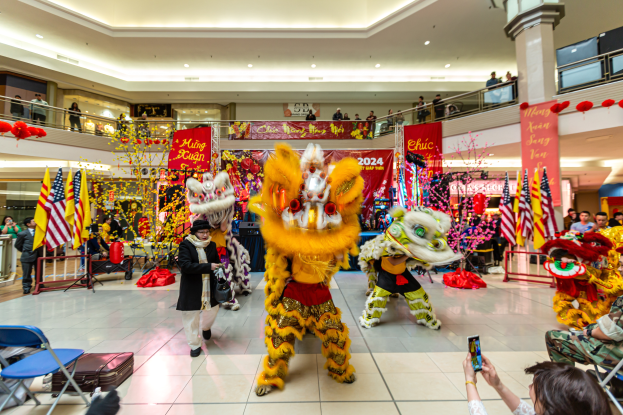 Ein lebendiger Chinese New Year-Feier in einem Einkaufszentrum mit tanzenden Menschen in Kostümen und anderen, die auf Stühlen sitzen und mobile Telefone halten, umgeben von Fahnen, Bannern, Dekorationen und architektonischen Elementen mit einem Wasserzeichen unten.