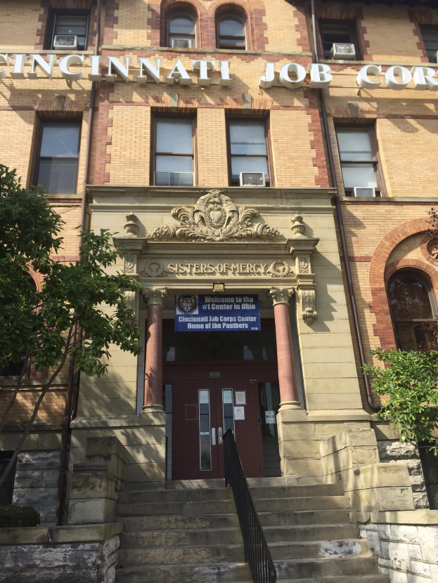 Exterior view of the Cincinnati Job Corps Headquarters building in Cincinnati, Ohio, featuring steps with railings, a name board, pillars, windows, doors, surrounding trees, and a visible sky in the background.