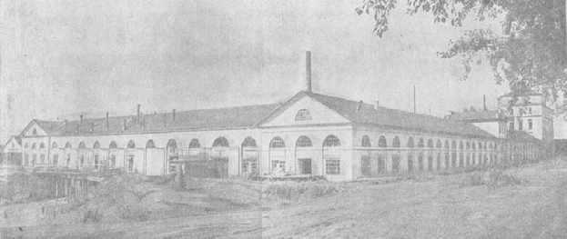 Schwarz-weiß-Foto eines großen mehrstöckigen Gebäudes mit zahlreichen Fenstern, umgeben von Grün, unter einem klaren Himmel, mit dem Text "1892 – das erste Fabrikgebäude in der Stadt" unten.