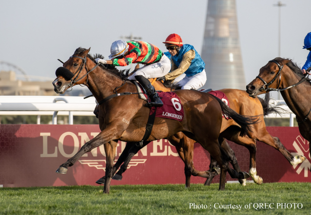 Drei Jockeys auf Pferden, die auf einem grasbedeckten Parcours mit einer Texttafel und einem Turm im Hintergrund unter einem klaren blauen Himmel galoppieren.