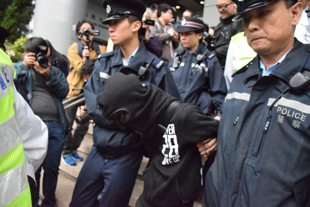 Eine Gruppe von Polizisten steht vor einer Menschenmenge, einige tragen Mützen und halten Kameras, mit einem Gebäude mit Säulen und Lampen im Hintergrund und Bäumen auf der linken Seite.