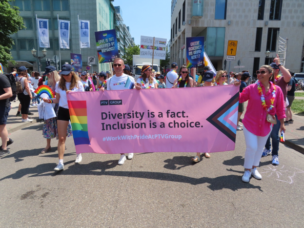 Eine Gruppe von Menschen, die eine Straße entlanggehen und ein Banner halten, auf dem steht "Diversität ist eine Tatsache, Inklusion ist eine Wahl", wobei einige Mützen und Schutzbrillen tragen und andere Banner halten, vor dem Hintergrund von Gebäuden, Bäumen und einem klaren blauen Himmel.