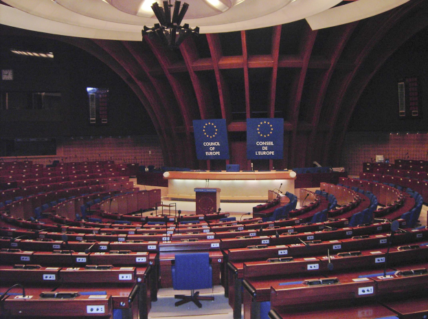 Großer halbrunder Konferenzraum mit Tischen und Stühlen, einem zentralen Podium und Wandtafeln mit Texten, der von Deckenleuchten in Brüssels Europäisches Parlament erhellt wird.