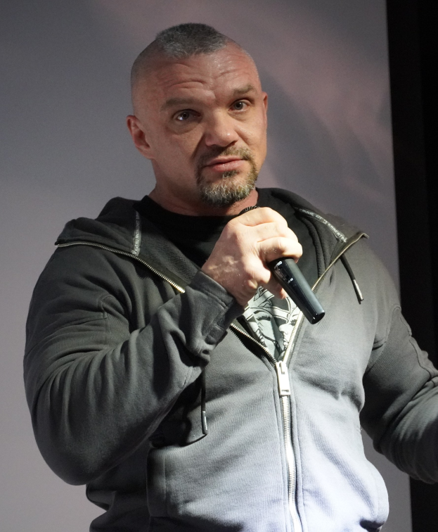 Ein Mann in einer schwarzen Kapuzenjacke und einem schwarzen Anzug mit weißem Hemd hält ein Mikrofon und steht selbstbewusst vor einer Wand, der Mann sieht aus wie ein Komiker mit zurückgekämmtem Haar.