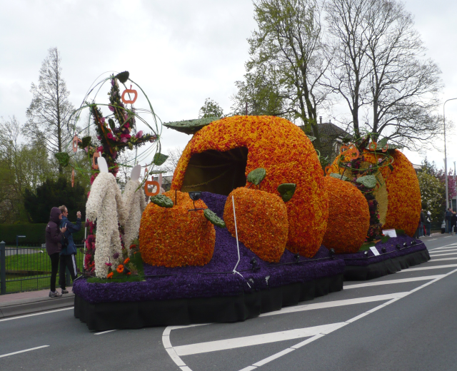 Schauwagen mit Kürbissen in einem Umzug, umgeben von Zuschauern auf der linken Seite, mit Bäumen, Gebäuden und Laternenmasten im Hintergrund unter einem klaren blauen Himmel.