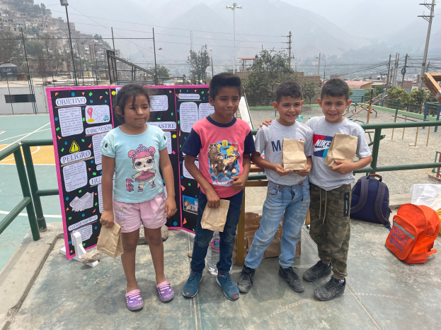 Eine Gruppe von Kindern steht auf einem Basketballplatz, jeder hält einen Papiertüte, mit Vorräten rechts daneben und einer Texttafel links, mit Zaun, Pfosten, Straßenlaternen, Bäumen, Gebäuden und Hügeln im Hintergrund.