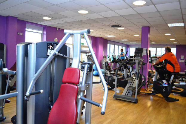 Ein Fitnessstudio mit verschiedenen Geräten und einigen Personen, die trainieren, mit einer Decke und Lampen oben