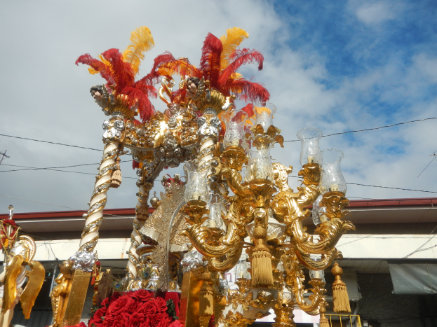 Ein prächtiger gold- und rotfarbener Festwagen, geschmückt mit Blumen und anderen Dekorationen bei einem Karnevalsumzug, mit einem Gebäude, Strommasten mit Drähten und einem bewölkten Himmel im Hintergrund.
