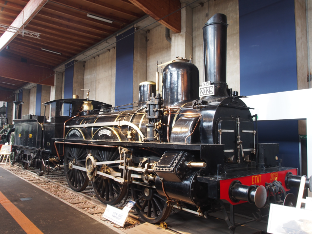 Eine große, alte Dampflokomotive in einem Museums-Setting umgeben von Eisenbahnschienen, Informations-Tafeln und anderen Ausstellungen, mit einer beleuchteten Decke und Wand im Hintergrund.
