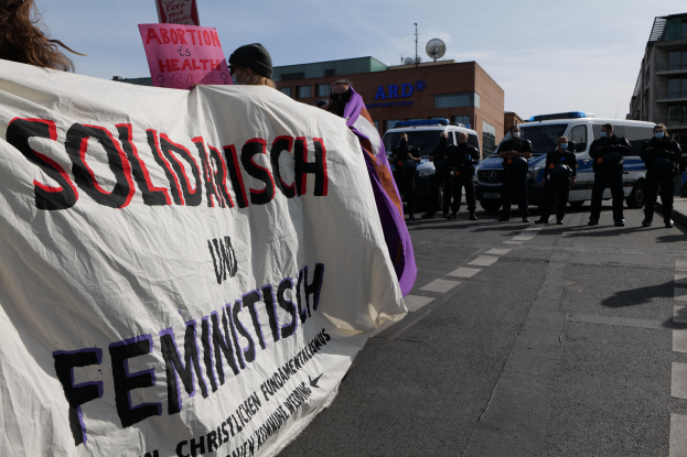 Eine Gruppe von Menschen marschiert auf der Straße und hält ein Transparent mit der Aufschrift 'Solidarität und Feminismus', im Hintergrund sind geparkte Fahrzeuge und Gebäude zu sehen.