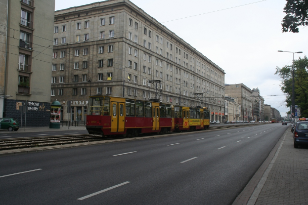 Eine Stadtansicht mit Gebäuden, Fahrzeugen, einem Zug, Straßenlaternen, Masten, Kabeln, einer Straße, Bäumen, Fenstern, Geschäften und einer bewölkten Himmel.