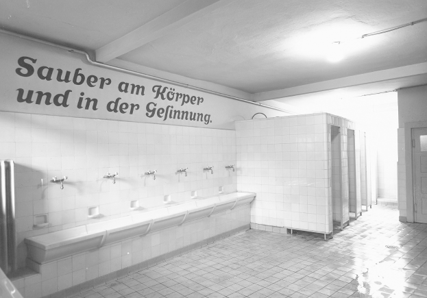 Schwarz-weiß-Foto einer öffentlichen Toilette mit Fliesenboden, Text an einer Wand, Wasserhähnen, einer Tür und einer Deckenlampe.
