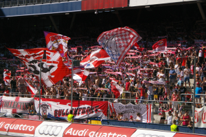 Eine große Menschenmenge in einem Stadion mit Fahnen, Bannern, einem Metallzaun, einem Schild, einem Bildschirm und Deckenleuchten.