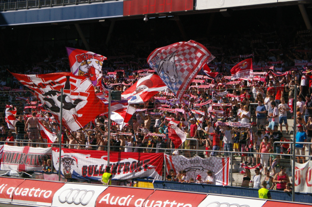Eine große Menschenmenge in einem Stadion mit Fahnen, Bannern, einem Metallzaun, einem Schild, einem Bildschirm und Deckenleuchten.