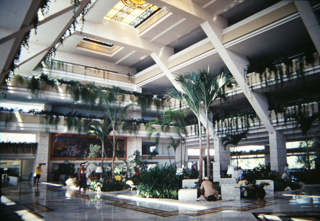 Eine geräumige Hotellobby mit einem hohen Atrium, das Menschen, Grünpflanzen, Säulen und Beleuchtung unter einer beleuchteten Decke zeigt.
