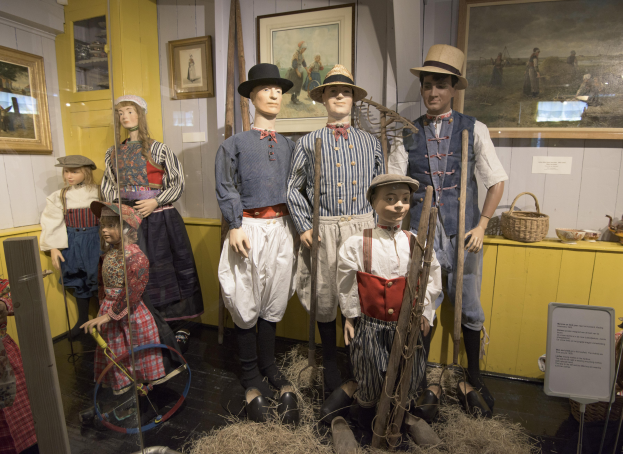 Eine Gruppe von Schaufensterpuppen in historischer Kleidung steht zusammen in einem Raum mit trockenem Gras auf dem Boden, einem Textbrett, an der Wand montierten Fotorahmen, einem Schrank mit Gegenst├Ąnden und einem Korb und anderen Gegenst├Ąnden auf einer Plattform.
