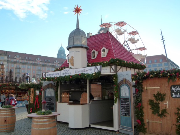 Ein kleines Gebäude mit einem roten Dach und einem Riesenrad davor, umgeben von Festdekorationen und Menschen, mit Gebäuden, Bäumen und einem bewölkten Himmel im Hintergrund während des Oktoberfests in München, Deutschland.