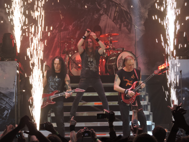 Eine Gruppe von Menschen, die auf einer Bühne mit Gitarren, einem Mikrofon und Trommeln spielt, während das Publikum das Konzert mit Handys aufnimmt.