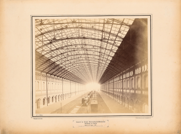 Schwarzes und weißes Foto eines Bahnhofs mit einem Zug auf den Schienen, Gebäuden mit Fenstern und einem Dach, das von Metallpfosten gestützt wird; unten ist Text sichtbar.