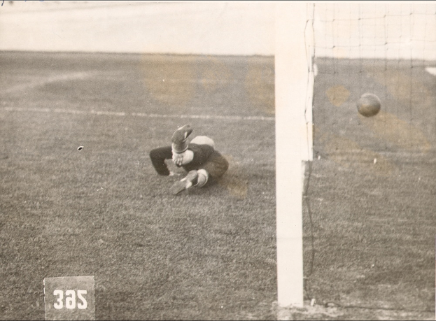 Ein Schwarz-Weiß-Foto eines Fußballspielers, der einen Ball vor einem Torpfosten mit Netz auf der rechten Seite und Text unten fängt.