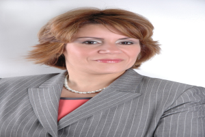 Eine professionelle Frau in einem blauen Anzug mit einem weißen Hemd und einer gemusterten Krawatte posiert selbstbewusst gegen einen weißen Hintergrund, ihre Haltung aufrecht und ihr Blick direkt.