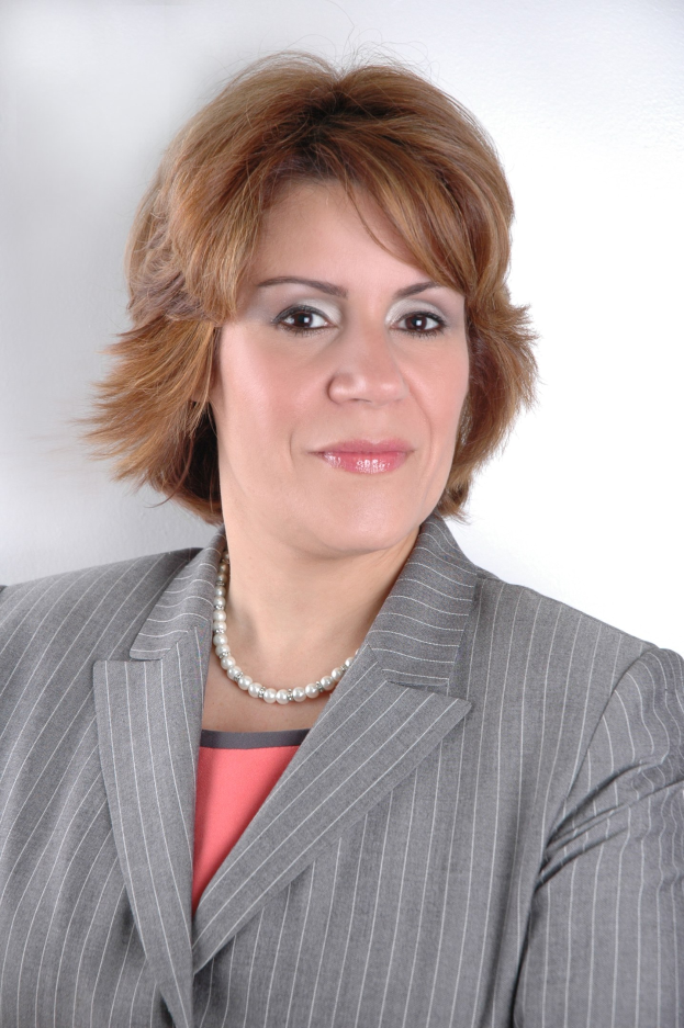 Eine professionelle Frau in einem blauen Anzug mit einem weißen Hemd und einer gemusterten Krawatte posiert selbstbewusst gegen einen weißen Hintergrund, ihre Haltung aufrecht und ihr Blick direkt.