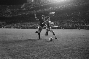 Zwei Männer spielen Fußball auf einem Feld in einem Stadion, umgeben von Zuschauern in den Rängen, beleuchtet von Flutlicht, in Schwarz-Weiß.