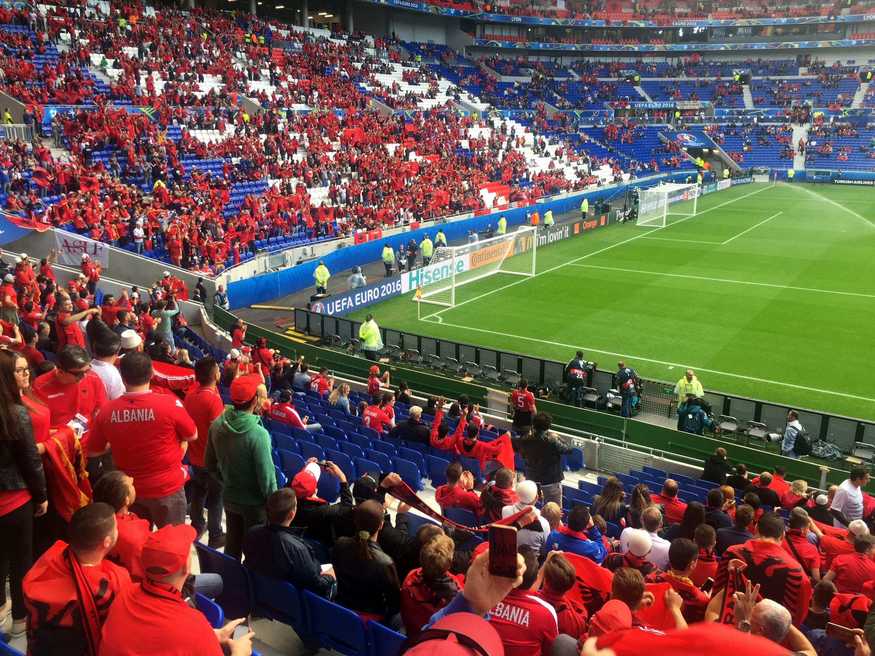Eine große Menschenmenge in einem Stadion schaut einem Fußballspiel zu, mit Menschen, die auf Stühlen sitzen und stehen, Bannern, einem Torpfosten und Deckenlichtern.