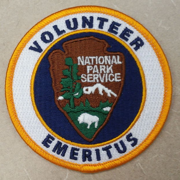 Ein rundes Abzeichen mit blauem Rand und weißem Hintergrund, das die fettgedruckte schwarze Schrift 'Volunteer National Park Service Emeritus' in der Mitte eines gelben Kreises zeigt.