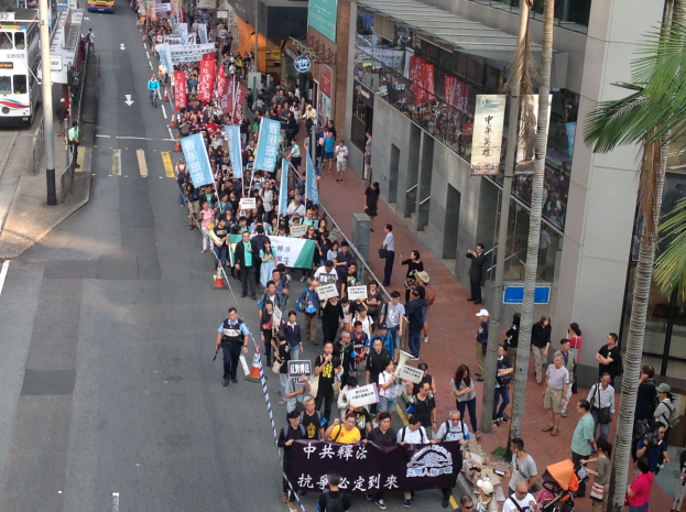 Eine große Gruppe von Menschen marschiert auf einer Straße in Hong Kong, hält Schilder und Plakate hoch, mit Bäumen, Glasfassaden, Fahrzeugen und Schildern auf beiden Seiten.