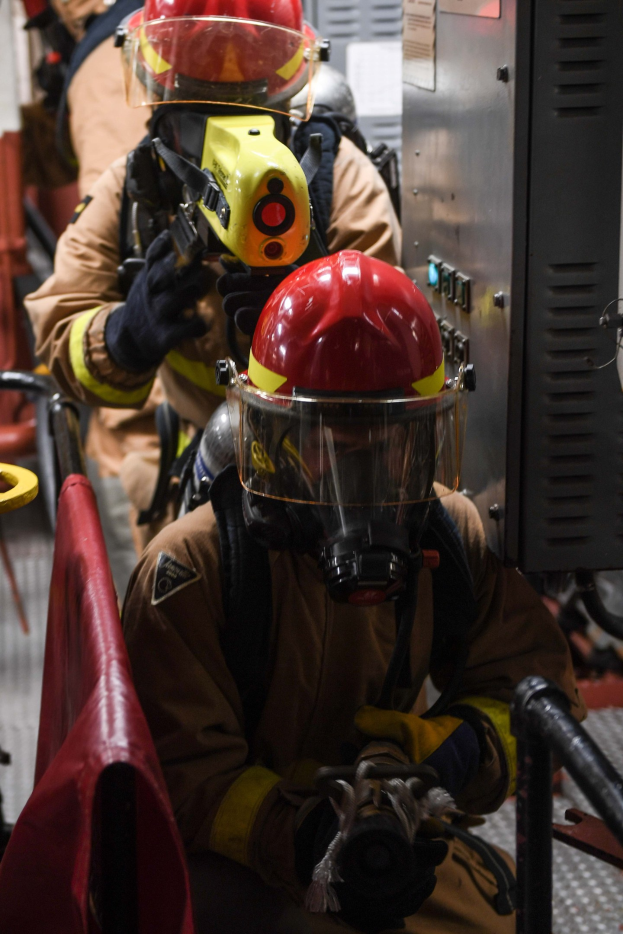 Feuerwehrleute in Schutzausrüstung mit einem Feuerlöscher, einem Metallobjekt und einem roten Tuch im Hintergrund.