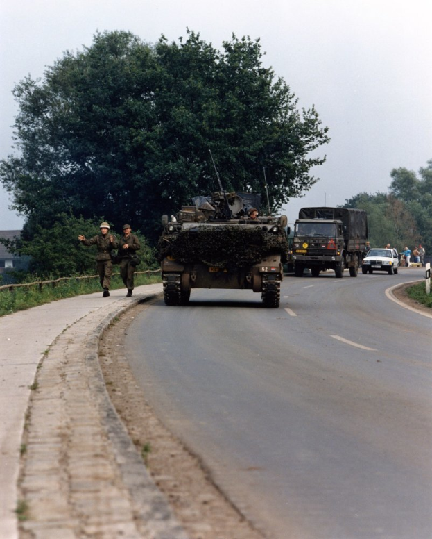 Ein Militärfahrzeug fährt auf einer Straße neben einem Wald, während uniformierte und helmtrugende Personen daneben gehen, während im Hintergrund Gebäude und ein klarer blauer Himmel zu sehen sind.
