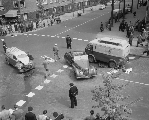 Schwarz-weiß-Szene eines Autounfalls an einer Straßenkreuzung mit einem Van, anderen Fahrzeugen und einer Gruppe von Menschen, die daneben stehen, mit Gebäuden, Bäumen, Pfählen und Texttafeln im Hintergrund.
