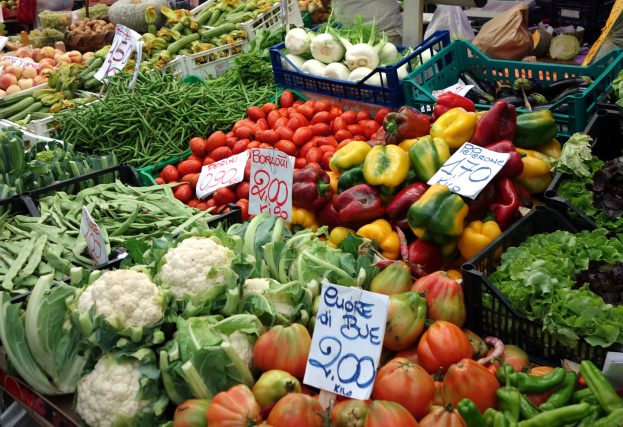 Ein belebter Bauernmarkt mit Körben voller Tomaten, Paprika, Blumenkohl, grünen Bohnen und anderen Gemüsen, jeweils mit Preisschildern, und einigen Menschen drumherum.