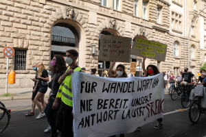 Eine Gruppe von Menschen, die auf einer Demonstration durch die Straße marschieren, einige halten Schilder und andere fahren Fahrräder, im Hintergrund ein Gebäude mit Fenstern, Bogengängen, Säulen und Skulpturen.