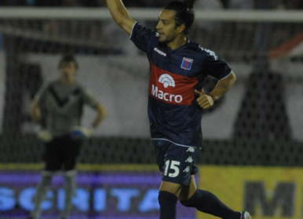 Ein Fußballspieler feiert nach einem Tor auf dem Boden, mit einer jubelnden Menge und einer Person im Hintergrund, alles vor einem unscharfen, energischen Hintergrund.