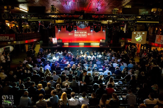 Eine große Zuschauermenge sitzt in Reihen von Stühlen, die einer hell erleuchteten Bühne mit Bildschirmen und Bannern gegenüberliegen und auf ein Online-Marketing-Event warten.