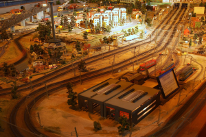 Eine detaillierte Miniatur-Modellbahnausstellung in einem Museum, die Gebäude, Bäume, Eisenbahnschienen, Fahrzeuge, Masten, Lichter, Schilder und andere Objekte umfasst, die eine belebte Stadtlandschaft nachahmen.