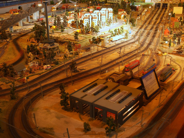Eine detaillierte Miniatur-Modellbahnausstellung in einem Museum, die Gebäude, Bäume, Eisenbahnschienen, Fahrzeuge, Masten, Lichter, Schilder und andere Objekte umfasst, die eine belebte Stadtlandschaft nachahmen.