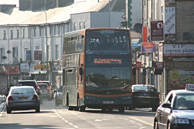 Eine Straße mit Autos und einem Bus im Vordergrund, mit Gebäuden im Hintergrund, die Wände, Fenster, Teller und Dächer aufweisen, sowie Plakate und Banner an den Wänden und einen Pfahl mit einer Straße Laterne.