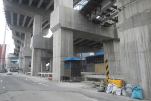 Große Betonstruktur im Bau über einer Straße mit darunterfahrenden Fahrzeugen, Folienabdeckungen und einem Schuppen auf dem Boden sowie Gebäuden im Hintergrund.