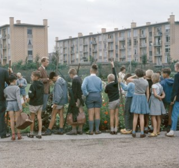 Kinder vor einem Gebäude mit Bäumen, Blumen und einem klaren Himmel, einige halten Schulranzen.