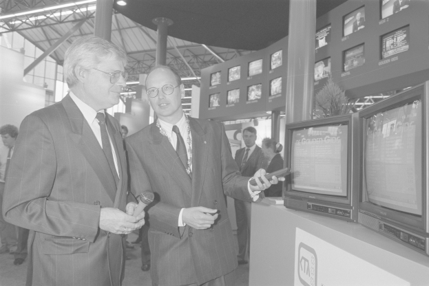 Zwei Männer in Anzügen stehen nebeneinander vor einem Fernseher und halten jeweils ein Mikrofon in der Hand, während im Hintergrund ein paar Menschen zu sehen sind. Das Bild ist in Schwarz-Weiß.