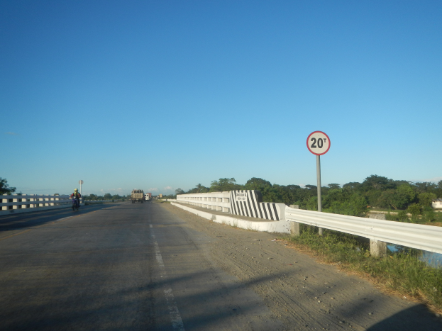 Eine Fahrbahn mit Fahrzeugen, ein Tempolimit-Schild, Gel"ander, Gras, B"ume und einen klaren blauen Himmel.