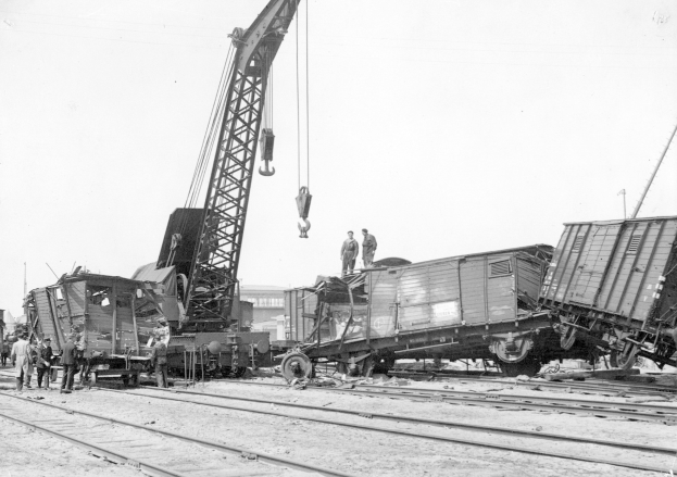 Schwarzes und weißes Bild einer Zugkatastrophe mit einem Kran, der einen Zugwagen auf die Schienen hebt, mehrere Menschen in der Nähe und im Hintergrund Gebäude und einen klaren Himmel.